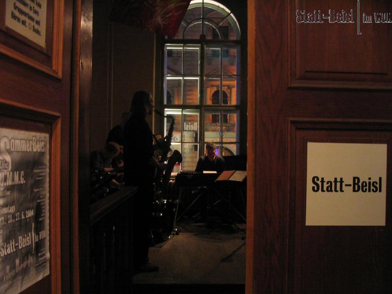 Links ein Türflügel mit einem SChild "Statt-Beisl", rechts im Hintergrund sitzen Gäste
