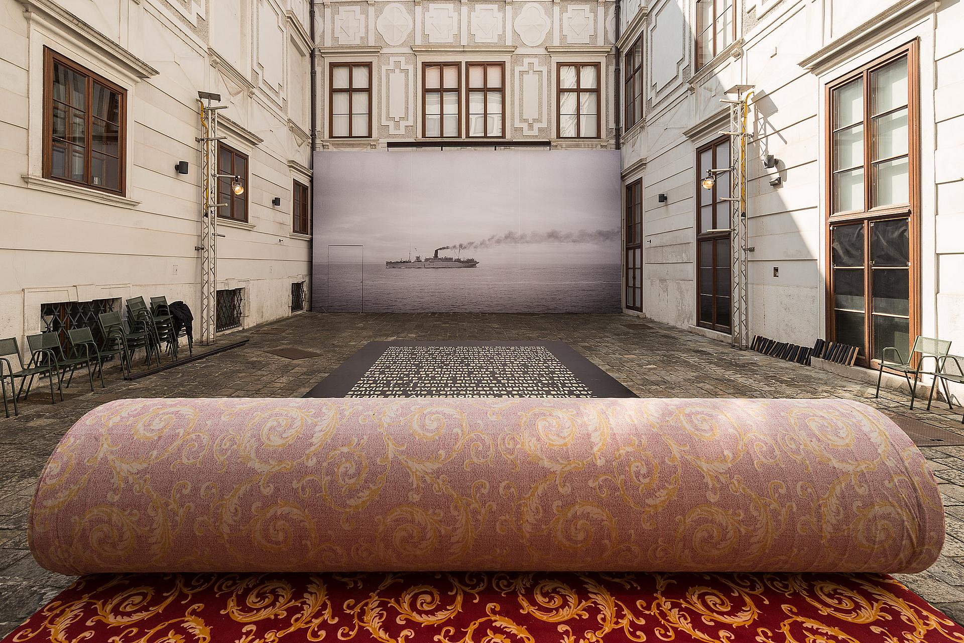 Innenhof mit aufgerolltem Teppeich im Zentrum.Vor dem teppich ist ein großes Plakat mit text auf dem Boden und an der hinteren Wand ist eine meterhohe schwaz weiß Fotografie eines Schiffes auf dem meer zu erkennen.
