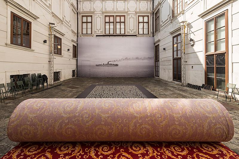 Innenhof mit aufgerolltem Teppeich im Zentrum.Vor dem teppich ist ein großes Plakat mit text auf dem Boden und an der hinteren Wand ist eine meterhohe schwaz weiß Fotografie eines Schiffes auf dem meer zu erkennen.