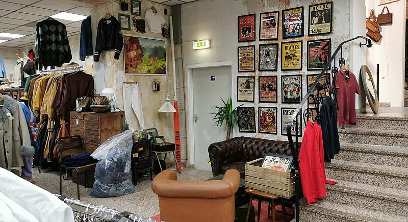 Bilder von verschiedenen Bands aus den 70ern und 80ern sind an einer Wand neben einer grauen Treppe zu sehen, während eine Couch davor und ein Notausgang daneben sind. Zusätzlich erkennt man links von der Tür ein paar Shirts und Jacken