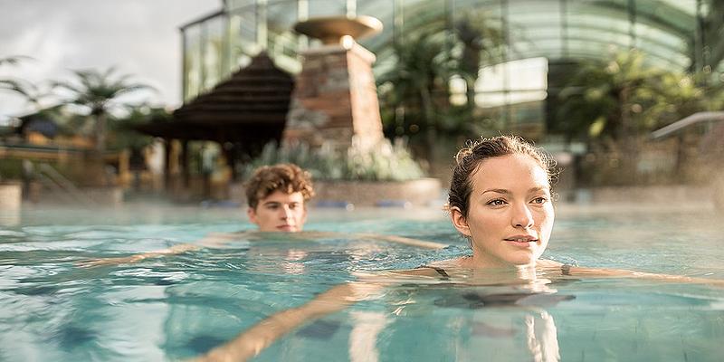 Frau schwimmt durch warmes Thermenwasser, dahinter ein Mann