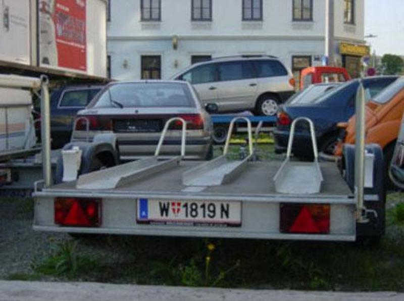 Leihanhänger mit offener Transportfläche, im Hintergrund: mehrere PKWs und ein Teil eines zweistöckigen Hauses sichtbar