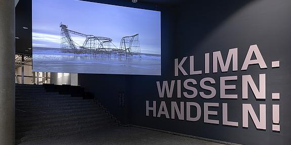 Filminstallation von Nikolaus Geyrhalter im Außenbereich der Dauerausstellung "Klima. Wissen. Handeln!" Auf der Wand steht in großen Lettern der Titel der Ausstellung.