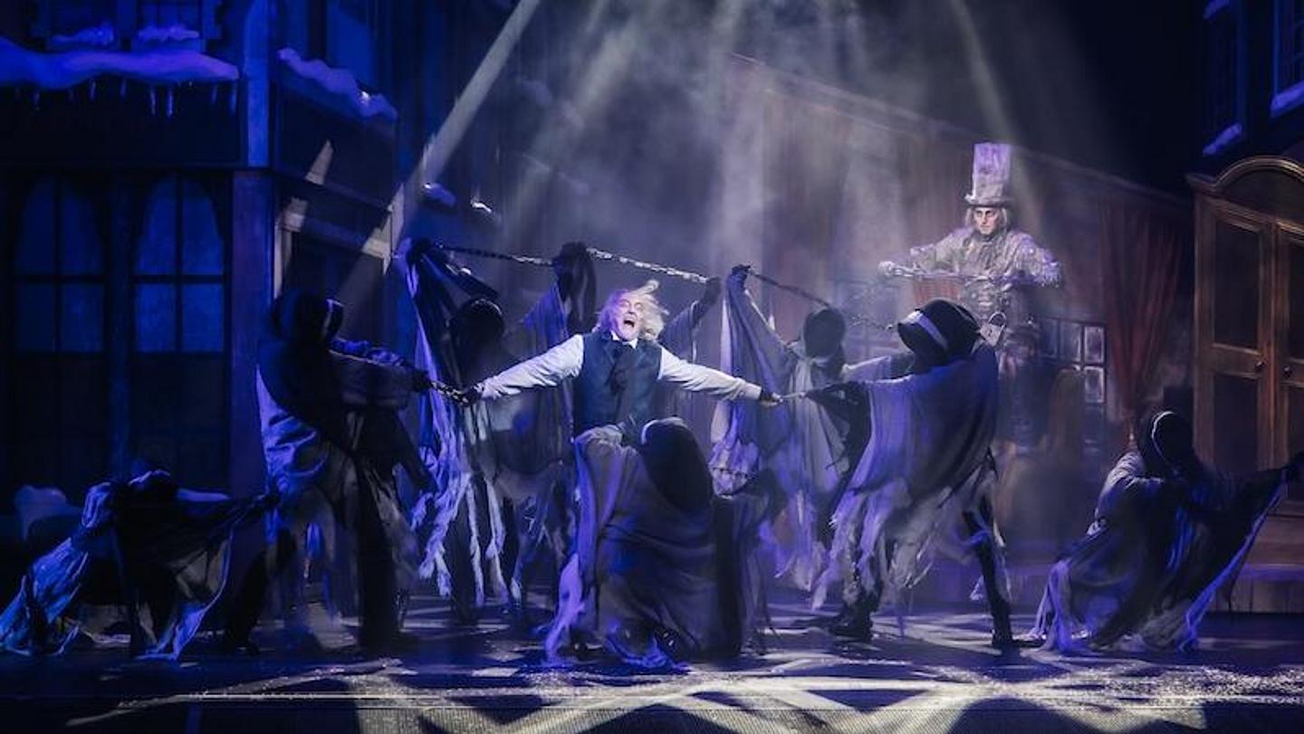 Musicalszene mit Ebenezer Scrooge und drei Weihnachtsgeistern vor winterlicher Kulisse- festlich beleuchtete Bühne mit weihnachtlicher Stimmung.