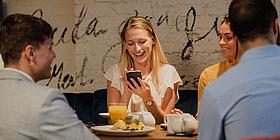 junge frau sitzt mit anderen am Tisch uns schaut lachend in ein Mobile