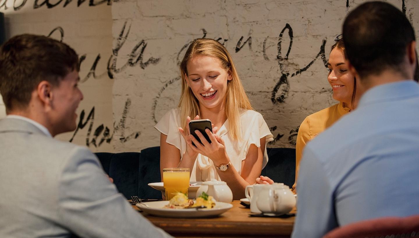 junge frau sitzt mit anderen am Tisch uns schaut lachend in ein Mobile