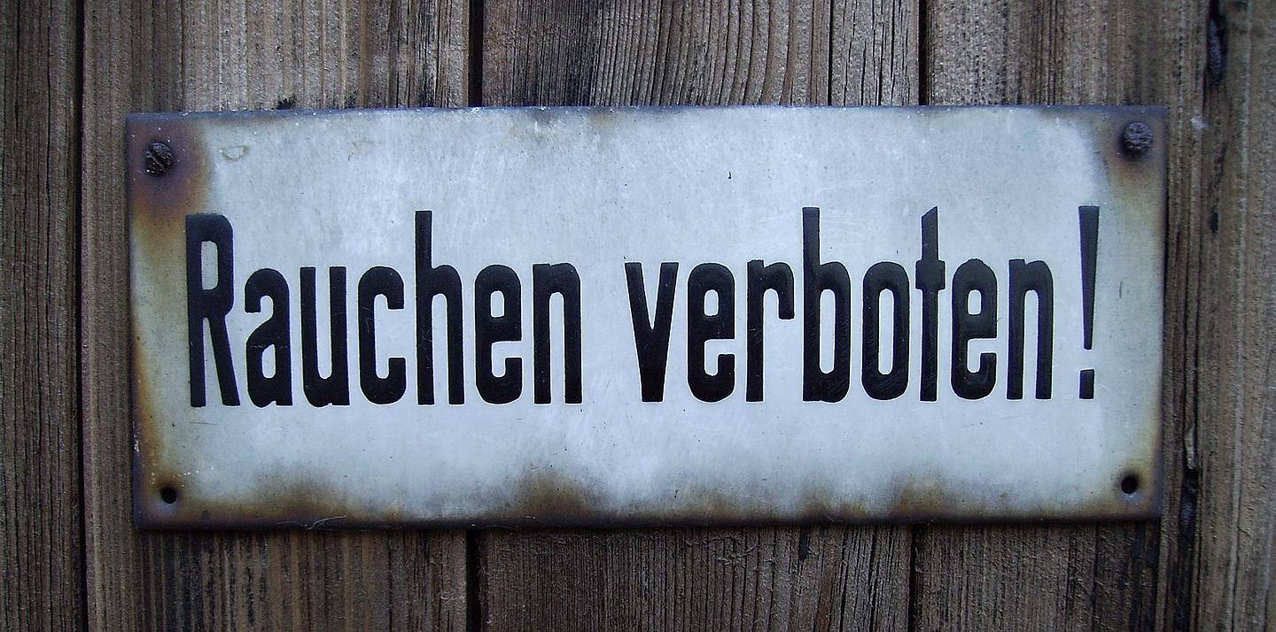 Weißes Metallschild mit der Aufschrift "Rauchen verboten!" auf hölzernem Hintergrund
