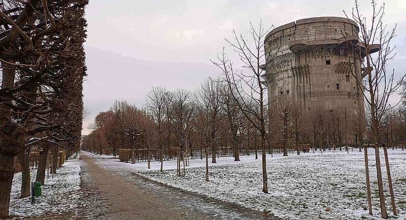 Ein gerader Weg führt durch eine Allee während nebenbei ein imposanter Flakturm auf einer Wiese steht