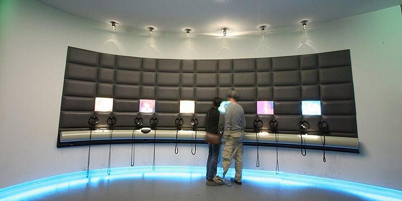 2 Personen vor 6 Bildschirmen die von einer gepolsterten Wand umgeben sind
