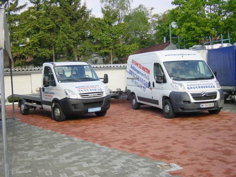 Zwei Kleintransporter, beide weiß mit roter und blauer Firmenaufschrift, der eine mit offener Transportfläche.