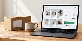 Laptop mit Online-Shop-Ansicht, links davon: Kaffeetasse und Schachtel 