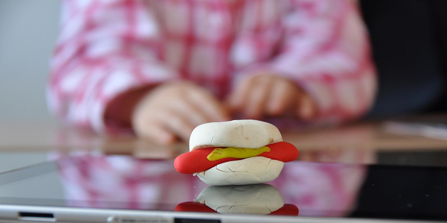 Kleiner Junge in weiß-rosa karriertem Hemd greift nach einer Hotdog-Semmel aus Knetmasse, man sieht nur angeschnittenen Oberkörper und Hände