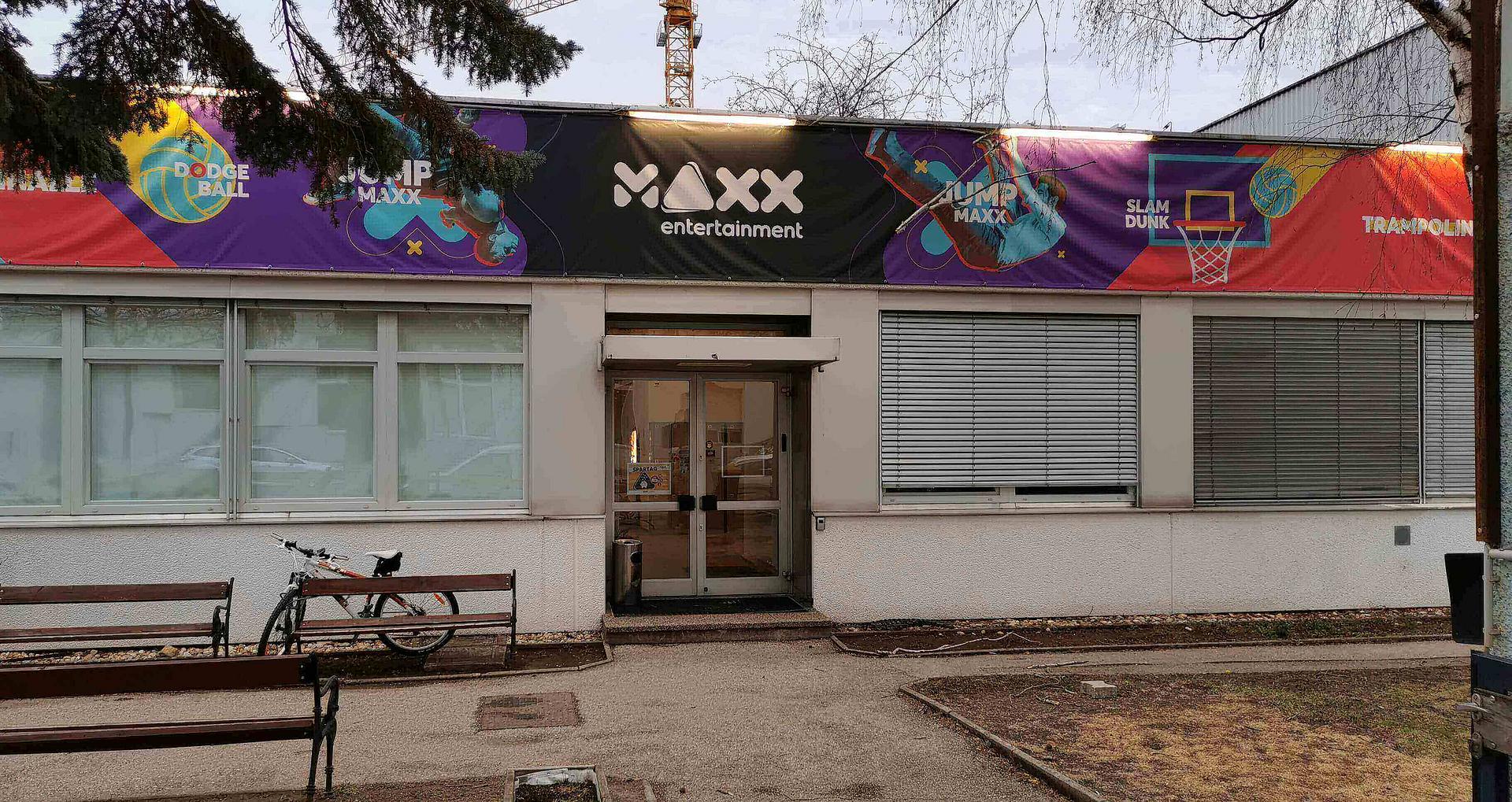 Die bunte Vorderseite eines Gebäude ist zu sehen; auf schwarz steht in weißer Schrift "MAXX". Neben dem Eingang steht ein Fahhrad hinter einer Sitzbank.