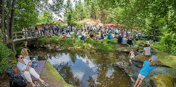 Schrammelklangfestival Grünes Waldstück mit einem kleinen See, bei dem einige Menschen sitzen.