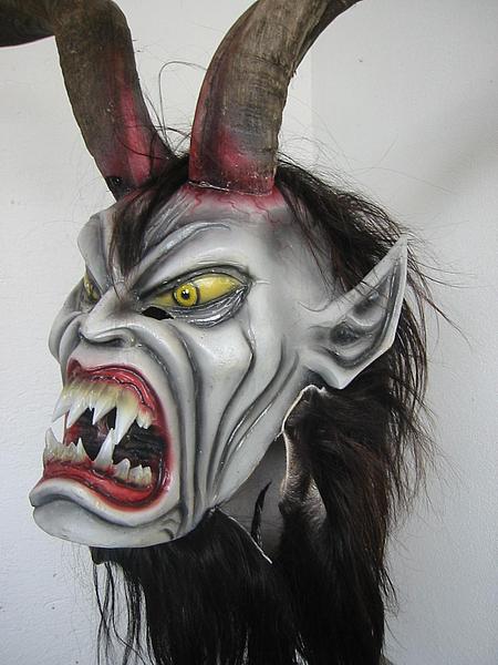 Zu sehen ist eine originale Krampusmaske mit einem weißen Gesicht, einen großen Mund mit spitzen Gangzähnen, gruseligen gelben Augen, spitzen Ohren und Hörnern.
