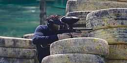 Einige Autoreifen, die aufeinander liegen und mit Farbe und Dreck versehen sind. Ein Mann, der sich hinter den Reifen versteckt und mit seiner Paintball-Waffe auf etwas zielt. 