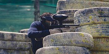 Einige Autoreifen, die aufeinander liegen und mit Farbe und Dreck versehen sind. Ein Mann, der sich hinter den Reifen versteckt und mit seiner Paintball-Waffe auf etwas zielt. 