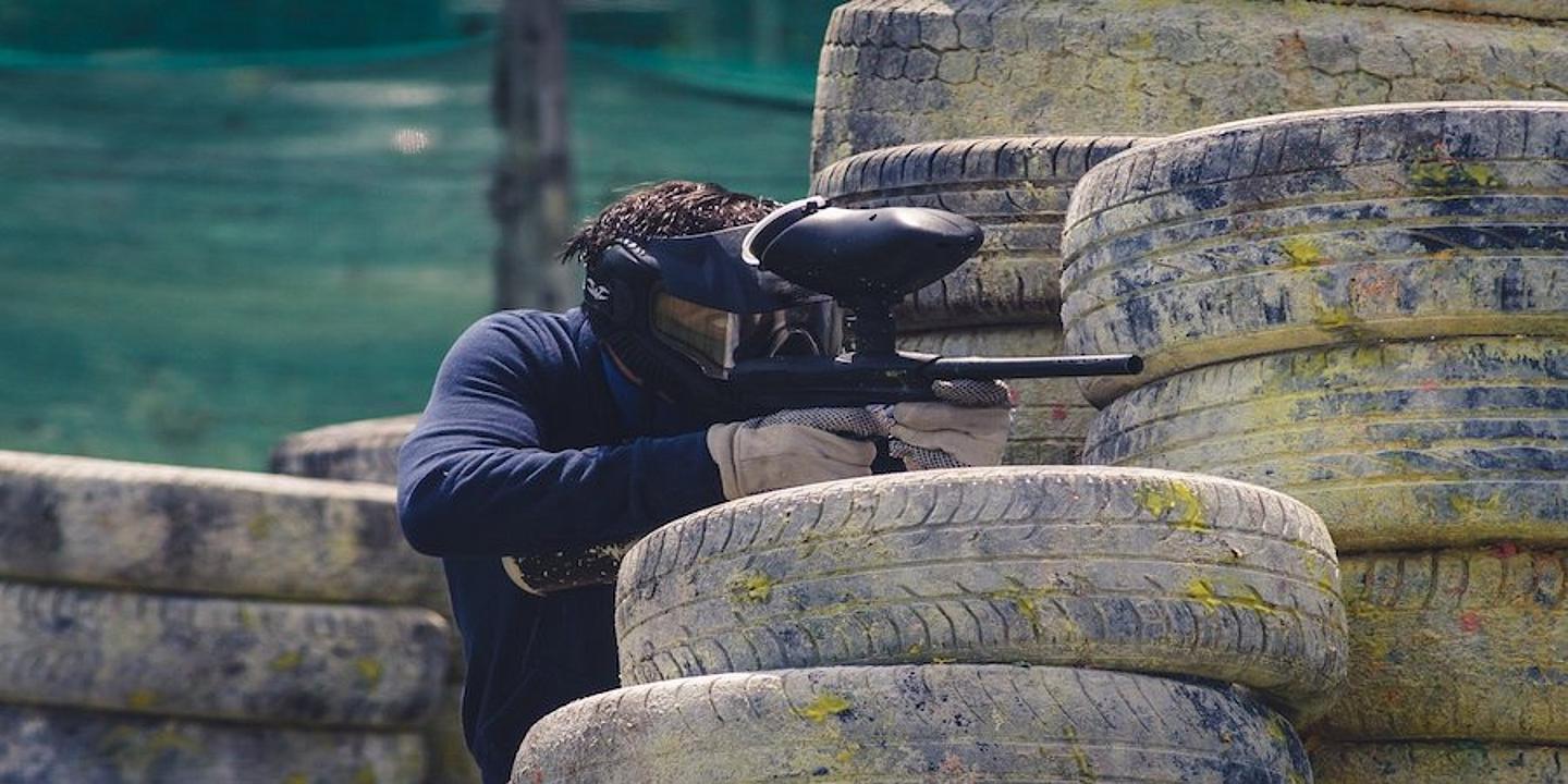 Einige Autoreifen, die aufeinander liegen und mit Farbe und Dreck versehen sind. Ein Mann, der sich hinter den Reifen versteckt und mit seiner Paintball-Waffe auf etwas zielt. 