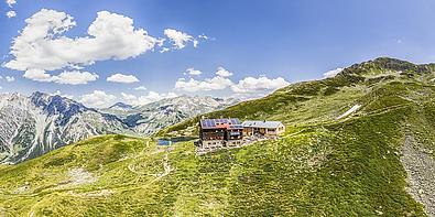 Kleine Almhütte auf grüner Wiese, im Hintergrund verschneiter Berg