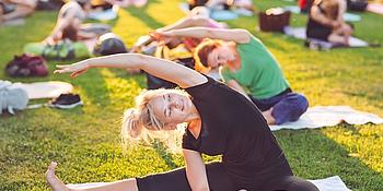 Bewegt im Park Symbolbild: Im Vordergrund Frau auf Yogamatte sitzend in Stretch-Position auf einer Wiese, im Hintergrund weitere Menschen in derselben Position