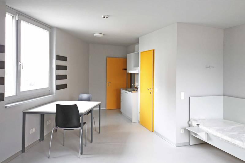 Single Appartement im Studentenwohnheim base 11. Raum in hellem weiß gehalten. Möbel weiß. Türen in dottergelb. Weißes Einzelbett. Schreibtisch beim Fenster mit zwei scharzen Stühlen.