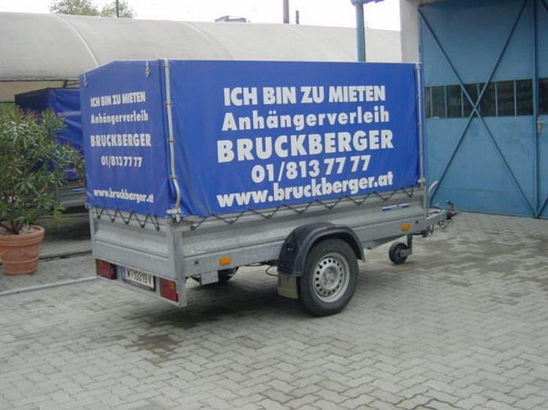Kastenförmiger Leihanhänger mit blauer Plane bespannt, drauf steht in weißer Schrift "Ich bin zu mieten Anhängerverleih Bruckberger 01 813 77 77 www.bruckberger.at