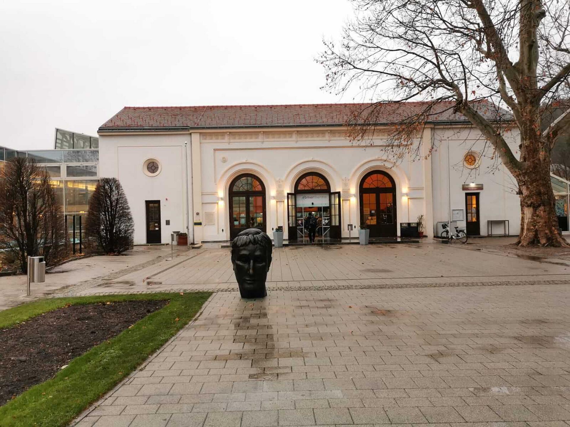 Ein großer asphaltierter Platz vor der Römertherme in Baden, während die Statue eines oxidiertetn Kaiserkopfes aus Bronze vor dem Gebäude steht.