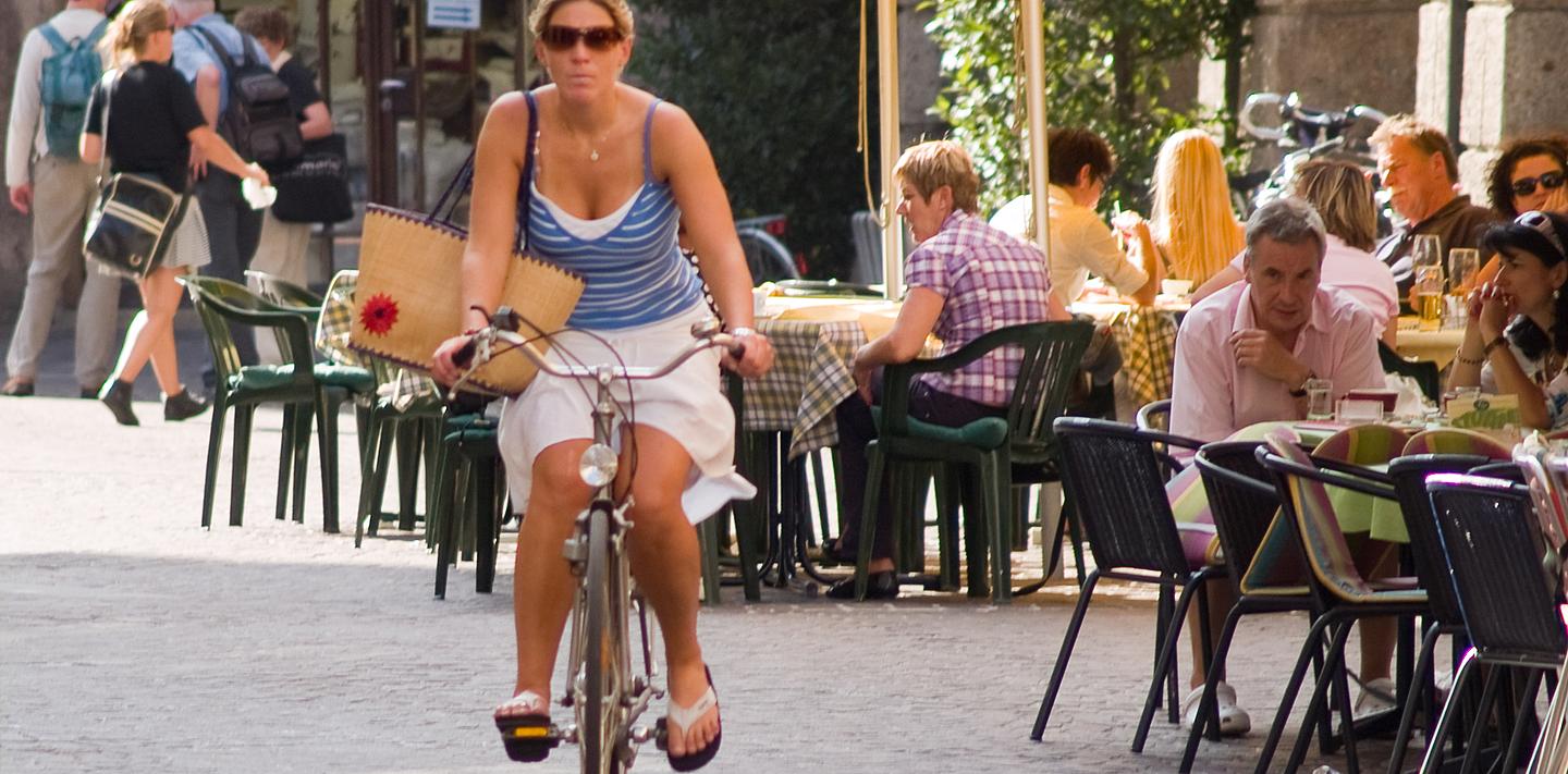Frau am Fahrrad in der Stadt dahinter Leute im Gastgarten