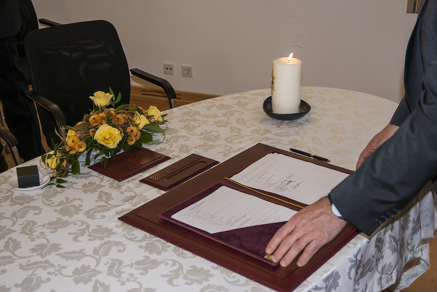 Eheschließund auf dem Standesamt: Auf einem Tisch mit weißer Tischdenke, Blumenkranz und einer brennenden Kerze sieht man die Trauurkunde