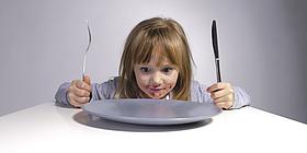Blondes Mädchen, das vor einem leeren Teller sitzt und Messer und Gabel in ihren Händen hält
