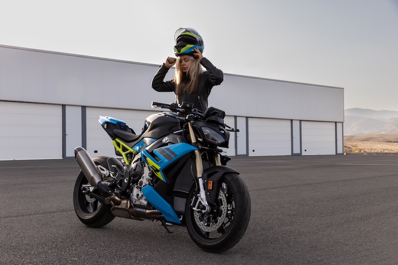 BMW S 1000 R 2025 – sportlicher Roadster stehend mit Frau, die Helm aufsetzt vor Halle, mit 165 PS; 999-ccm-Vierzylinder und Assistenzsystemen