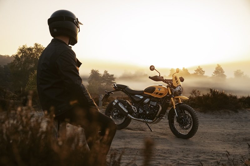 ﻿﻿Triumph Scrambler 400 XC Modelljahr 2025 – Fahrer schaut auf Retro-Bike stehend im Sonnenuntergang mit 398 ccm, 40 PS und Adventure-Look für Stadt und Land
